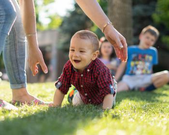 Smiling toddler crawling on grass, being supported by an adult