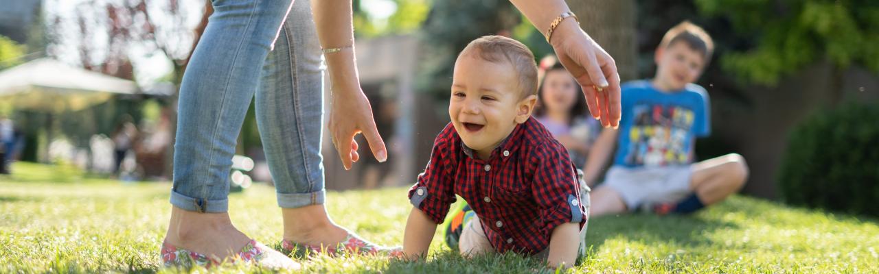 Smiling toddler crawling on grass, being supported by an adult